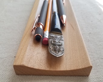 May include: A wooden pen holder with a curved edge, holding a black pen, a rose gold pen, a yellow pencil, and a silver letter opener with an ornate design.