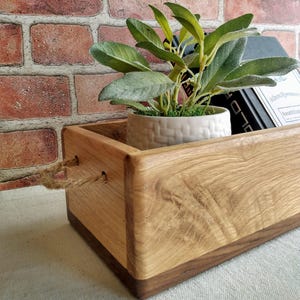 May include: A wooden box with rope handles, a white ceramic planter with a small plant, and a black book with the title "John Brown's Body" on the top. The box is made of light wood with a darker wood trim.