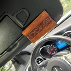 May include: A rectangular wooden car visor accessory with a rich, reddish-brown grain pattern. The accessory is mounted on a black car visor, with a portion of the car's interior, including the steering wheel and dashboard, visible in the background.