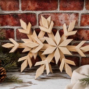 Puede incluir: Dos adornos de copo de nieve superpuestos hechos de madera natural de color claro. Cada copo de nieve presenta un diseño geométrico de estrella de múltiples puntas con segmentos angulares afilados que irradian desde un punto central.
