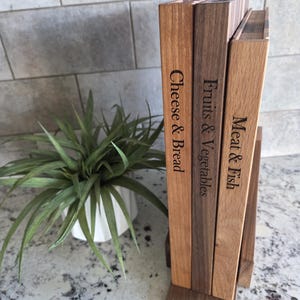 May include: Wooden cutting board set with engraved labels for Cheese & Bread, Fruits & Vegetables, and Meat & Fish. The boards are various shades of brown and are displayed upright in a wooden stand. A green plant in a white pot is in the background.
