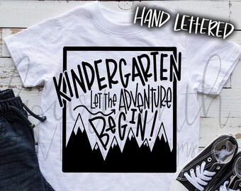 May include: White t-shirt with black text that reads "Kindergarten Let the Adventure Begin!" inside a black square with mountain peaks. The shirt is laid out on a white wood surface with a pair of black canvas shoes and blue denim shorts.