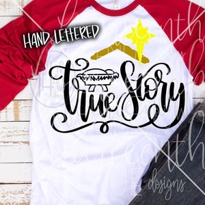 May include: A white long-sleeved shirt with a red raglan sleeve. The shirt has a black and gold design that says "Hand Lettered True Story" with a manger and a star.