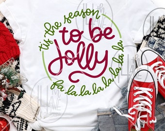 Puede incluir: Camiseta blanca con un círculo verde y el texto "'tis the season to be jolly la la la la la la la la" en letras rojas y verdes.