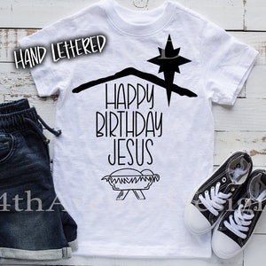 May include: White t-shirt with black text that reads "Happy Birthday Jesus" with a star and manger graphic. The shirt is on a wooden surface with a pair of black sneakers and blue denim shorts.