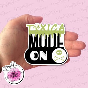 May include: A black and green sticker with the text "TOXICA MODE ON" in white lettering. A skull and crossbones symbol is in the lower right corner of the sticker.