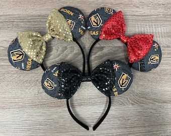 Puede incluir: Tres pares de orejas de ratón negras con una diadema. Cada par tiene un lazo de color diferente: dorado, rojo y negro. Las orejas están decoradas con un logotipo de los Vegas Golden Knights y las palabras "Vegas Born" y "Vegas Golden Knights".