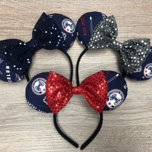 May include: Five headbands with mouse ears. The ears are navy blue with a white and red logo and a bow. The bows are navy blue with sequins, silver with sequins, and red with sequins. The headbands are black.