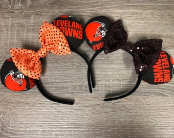 May include: A pair of black headband mouse ears with orange and black bows. The ears are decorated with a Cleveland Browns logo and the words "Cleveland Browns".