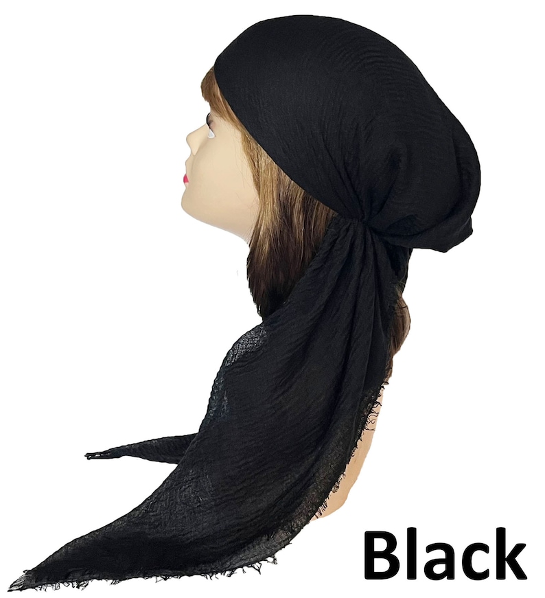 Op de afbeelding: Een effen zwarte hoofddoek met een gekreukte, getextureerde stofafwerking. De hoofddoek is aan de zijkant geknoopt en heeft lange, vloeiende uiteinden met gerafelde randen die naar beneden vallen. Het woord Black staat onderaan in vetgedrukt.