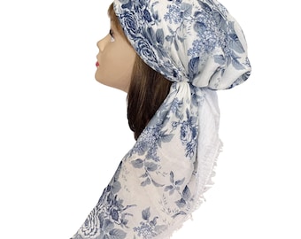 Op de afbeelding: Een hoofddoek met een witte basis en een blauw bloemenpatroon, vastgeknoopt aan de achterkant van het hoofd. De stof toont gedetailleerde illustraties van rozen en bladeren, met een fijne kanten rand langs de zijkanten voor een zachte uitstraling.