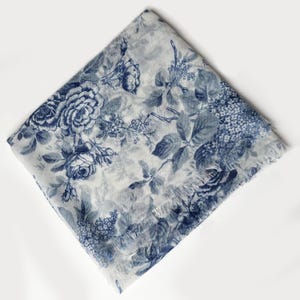 May include: A square scarf featuring a white base with a detailed blue floral print. The pattern displays various roses, leaves, and small blossoms across the fabric. The edges of the scarf are finished with a delicate, frayed fringe detail.