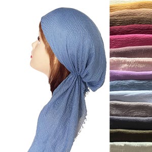 May include: A blue crinkled headscarf worn on a mannequin head, featuring a textured fabric with a loose drape and frayed edges. To the right, a vertical stack of folded headscarves is displayed in various colors including yellow, brown, pink, purple, and grey.