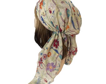 Op de afbeelding: Een lichtgewicht hoofddoek in een lichtbeige tint met een kleurrijk bloemen- en vogelpatroon. De stof toont diverse bloemen in tinten rood, paars en oranje, afgewisseld met blauwe vogels en dunne, kronkelende lijnen over het gehele oppervlak.