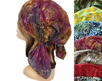 Puede incluir: Un pañuelo para la cabeza con un estampado floral estilo batik en tonos púrpura, magenta y dorado. La tela está fruncida en la parte posterior con un cierre de lazo. Una columna vertical a la derecha muestra variaciones de color en verde, amarillo, azul y negro.