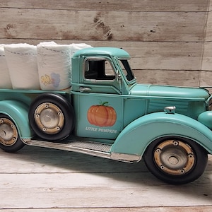 Könnte beinhalten: Ein türkisfarbener Vintage-Metall-Truck mit einer Kürbisgrafik und dem Text "Little Pumpkin" an der Seite. Die Ladefläche des Trucks enthält mehrere Rollen weißer Papiertücher. Der Truck hat schwarze Reifen und ein Used-Finish.