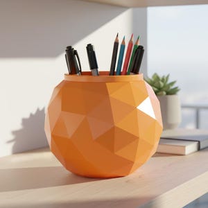 May include: An orange geometric pen holder filled with pens and pencils. The holder has a faceted design and sits on a wooden surface. The pens and pencils are black, blue, red, and green.