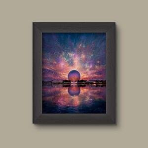 May include: Framed print of a colorful sunset over a lake, featuring a large geodesic sphere and fireworks. The sky transitions from purple to orange and pink, with the sphere reflecting in the water. The frame is black.