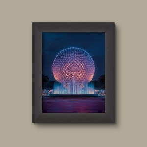 May include: Framed print of a large geodesic sphere with a geometric pattern, illuminated in shades of purple and orange. A fountain with blue water sprays in front of the sphere, with trees on either side. The image is framed in a black frame.