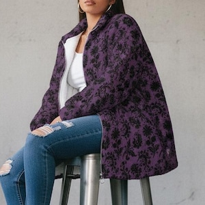 Könnte beinhalten: Eine violette Jacke mit schwarzem Blumenmuster, ein weißes Unterhemd, eine blaue Jeans mit Rissen an den Knien und schwarze Stiefeletten. Die Jacke hat einen Reißverschluss und einen hohen Kragen und wird über einem schlichten weißen Oberteil getragen.
