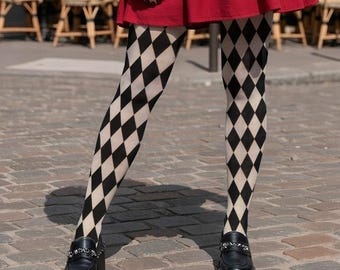 May include: A red V-neck dress with a tiered skirt, a black leather jacket worn over the shoulders, black and white diamond-patterned tights, black loafers, a small black crossbody bag, and black sunglasses, standing on a cobblestone street.
