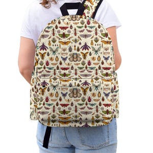 Op de afbeelding: Een rugzak met een all-over print van diverse insecten en vlinders in tinten rood, geel, groen, blauw en bruin op een crèmekleurige achtergrond. De tas heeft een voorvak met rits, zwarte schouderbanden en zwarte biezen.