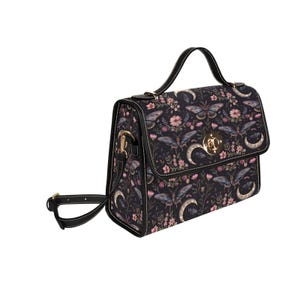 Könnte beinhalten: Eine schwarze Handtasche mit einem Blumen- und Schmetterlingsmuster. Die Tasche hat einen oberen Griff und einen abnehmbaren Schultergurt. Das Design umfasst rosa Blumen, Halbmonde und Schmetterlinge. Die Tasche hat einen goldfarbenen Verschluss.