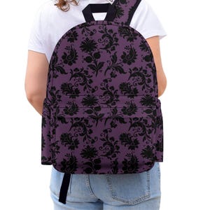 May include: A backpack featuring a dark purple base colour with a repeating black floral and paisley pattern. The bag includes a large main compartment, a front zippered pocket, and black shoulder straps, providing a functional storage solution.