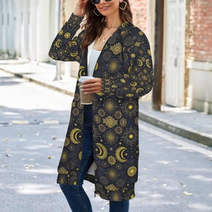 May include: A long-sleeved, knee-length zip-up hoodie in a dark navy blue fabric featuring an all-over gold and yellow celestial print of suns, moons, and stars. The garment is worn over a plain white top and dark blue denim jeans.