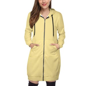 May include: A light yellow hooded sweatshirt dress with a full-length zipper and front pockets. The dress has long sleeves and a hood with black drawstrings. The model is wearing black thigh-high socks.