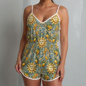 May include: A one-piece romper with a V-neck and spaghetti straps. The romper features a repeating pattern of smiling suns, lemons, and floral elements in yellow, blue, and white. The edges are trimmed with white piping.