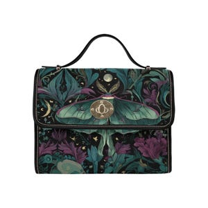 May include: A black handbag with a top handle and a flap closure. The bag features a detailed illustration of a Luna moth with teal wings, set against a backdrop of teal and purple foliage. The hardware is gold-toned.