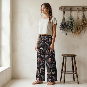 Op de afbeelding: Zwarte palazzo broek met een bloemen- en hemels patroon met vlinders, manen en bloemen. De broek heeft een wijd been ontwerp en een hoge taille. De achtergrond is zwart met roze, blauwe en witte accenten.