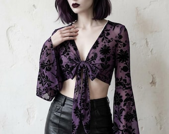 May include: A sheer purple crop top with long, flared sleeves and a tie-front closure. The top features a black floral pattern and a deep V-neckline. The garment is made of a lightweight fabric.