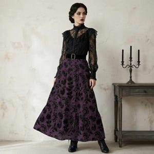 Op de afbeelding: Een zwarte kanten blouse met lange mouwen en een hoge kraag, gecombineerd met een paarse rok met bloemenprint en een zwarte riem. De rok is vloerlang. De outfit is in gotische stijl.