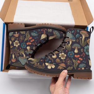 A hand holds up one of the mushroom-patterned lace-up boots outside of its open cardboard shoebox.