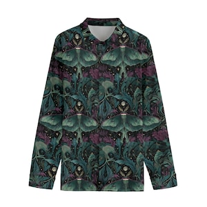 Puede incluir: Una camisa de manga larga con botones, con un patrón repetido de grandes polillas verdes, flores moradas y follaje verde oscuro sobre un fondo negro. La camisa tiene cuello y puños blancos. El diseño tiene una estética botánica vintage.