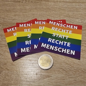 Könnte beinhalten: Mehrere Aufkleber in Regenbogenfarben mit dem Text "MENSCHEN RECHTE STATT RECHTE MENSCHEN". Eine Zwei-Euro-Münze liegt neben den Aufklebern. Die Aufkleber haben ein Regenbogenflaggen-Design.