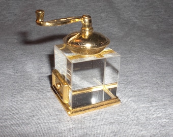 Puede incluir: Un molinillo de café en miniatura de color dorado con una base de vidrio transparente y un mango dorado. El molinillo está sobre una superficie gris.