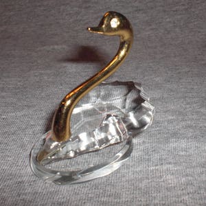 May include: A crystal swan ring holder with a gold-tone neck and head. The swan's head is tilted to the side and has a small, clear rhinestone in its eye.