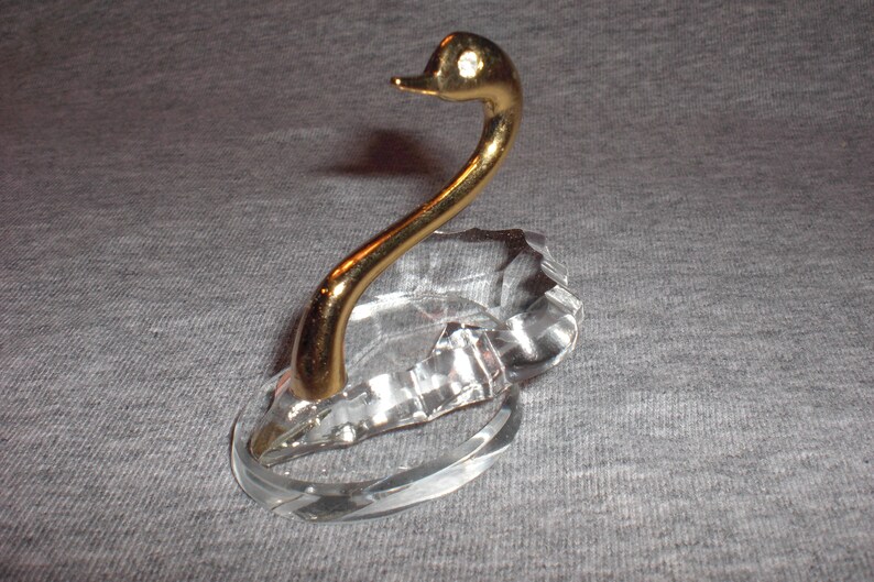May include: A crystal swan ring holder with a gold-tone neck and head. The swan's head is tilted to the side and has a small, clear rhinestone in its eye.