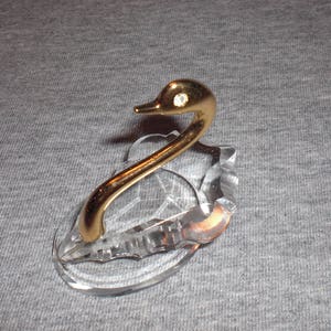 May include: A crystal swan figurine with a gold-tone neck and head. The swan has a single rhinestone on its head.