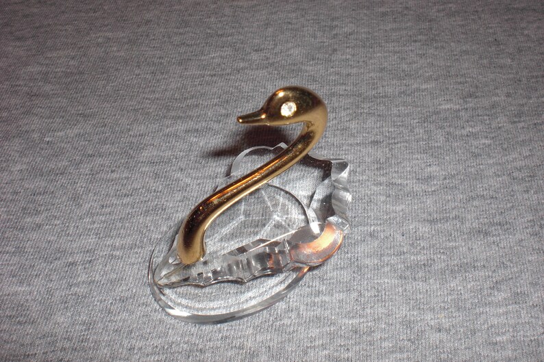 May include: A crystal swan figurine with a gold-tone neck and head. The swan has a single rhinestone on its head.