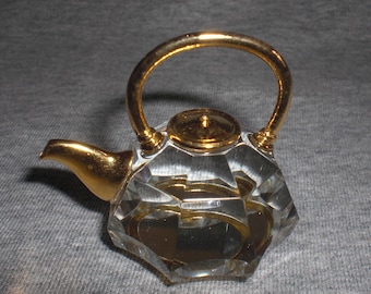 Puede incluir: Una tetera de cristal con asa y tapa chapadas en oro. La tetera es facetada y tiene un aspecto claro y vítreo.
