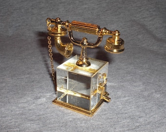 Puede incluir: Una figurita de teléfono rotatorio de estilo vintage de color dorado con una base de cristal transparente. El teléfono tiene una cadena unida al auricular.