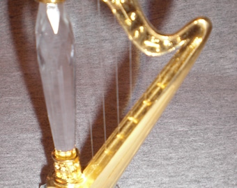 Puede incluir: Una figura decorativa de arpa con un marco dorado y una base de cristal transparente facetado. Las cuerdas del arpa son visibles, y el cuerpo es una combinación de oro y cristal transparente. Un artículo decorativo con temática de instrumentos musicales.