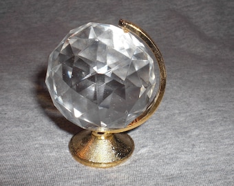 Puede incluir: Un pequeño globo de cristal con un soporte de metal dorado. El globo es facetado y transparente.