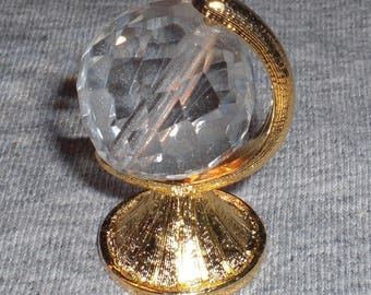 Puede incluir: Un pequeño globo de cristal sobre un soporte dorado. El globo es transparente y facetado, y el soporte tiene un diseño decorativo.