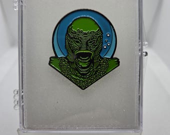 Peut inclure: Épingle en émail vert et noir représentant un design de créature, sur fond bleu et vert. L'épingle est présentée dans un étui en plastique transparent avec un intérieur blanc, pour les collectionneurs ou les fans de films de monstres.