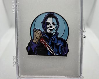 Peut inclure: Épingle en émail représentant un personnage de film d'horreur tenant un couteau ensanglanté. L'épingle a une palette de couleurs bleue et blanche, sur un fond bleu clair. L'épingle est enfermée dans un présentoir en plastique transparent.